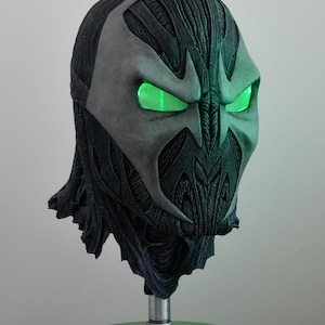 Puede incluir: Una máscara oscura y detallada con ojos verdes brillantes. La máscara tiene un diseño texturizado y orgánico con una placa facial gris y detalles intrincados. Los ojos brillan intensamente, agregando un elemento visual llamativo.