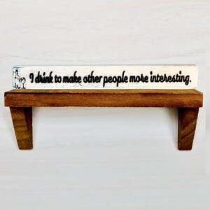 May include: A wooden shelf with a white sign that reads "I drink to make other people more interesting." The shelf is brown with two angled supports. The sign has a wine glass and bottle graphic.