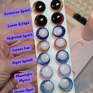 May include: A vertical display card featuring seven pairs of circular cabochon eyes with glittery iris designs. The labels include Evolution Spark, Lunar Eclispe, Nightfall Spark, Forest Fae, Aqua Splash, Moonlight Mystic, and Ocean Opal.