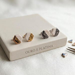 Könnte beinhalten: Zwei Paar sechseckige Ohrstecker mit Gold- und Silberakzenten. Die Ohrringe werden auf einem rechteckigen, cremefarbenen Block mit dem Text "OURO E PLATINA" präsentiert. Kleine Keramikfragmente und eine Pinzette sind ebenfalls zu sehen.