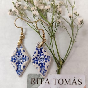 Pode incluir: Um par de brincos pendurados dourados com azulejos de cerâmica em forma de diamante com um design floral azul e branco. Os brincos são exibidos com flores brancas e um cartão de visita que diz "RITA TOMÁS Atelier Handmade Portugal."