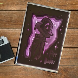 Könnte beinhalten: Ein lila und schwarzer gerahmter Druck einer Silhouette von Dobby, dem Hauselfen aus der Harry-Potter-Reihe. Der Druck hat einen 3D-Effekt und zeigt einen sternenübersäten Hintergrund. Der Text "Free Dobby" ist unten auf dem Druck gedruckt.