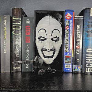 Könnte beinhalten: Auf einem schwarzen Regal stehen Bücher und VHS-Kassetten, darunter "CULT", "DER GEIST" und "GHOSTBUSTERS". Eine weiße Maske mit schwarzen Akzenten und eine kleine schwarze Pistole ergänzen das Arrangement, das ein Horror-Thema erzeugt.