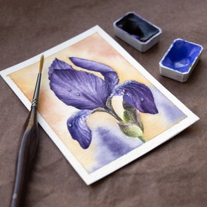 May include: A watercolor painting of a purple iris flower with water droplets on the petals. The painting is on a white paper background and there are two watercolor paint pans in the background.