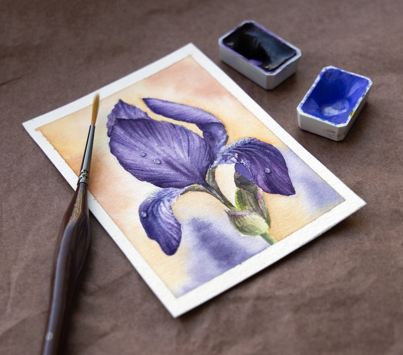 May include: A watercolor painting of a purple iris flower with water droplets on the petals. The painting is on a white paper background and there are two watercolor paint pans in the background.