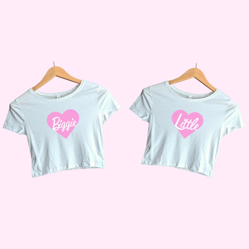 Pu&ograve; includere: Due crop top bianchi con cuori rosa e le parole "Biggie" e "Little" stampate su di essi. I top sono appesi a grucce di legno.