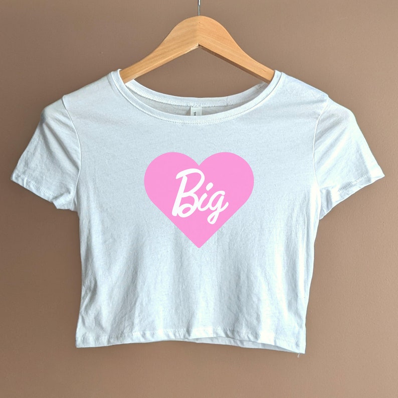 Pu&ograve; includere: Maglietta bianca corta con un cuore rosa su cui &egrave; scritto "Big" in corsivo bianco.