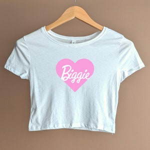 Pu&ograve; includere: Maglietta bianca corta con un grafico a forma di cuore rosa che dice "Biggie" in lettere bianche in corsivo.
