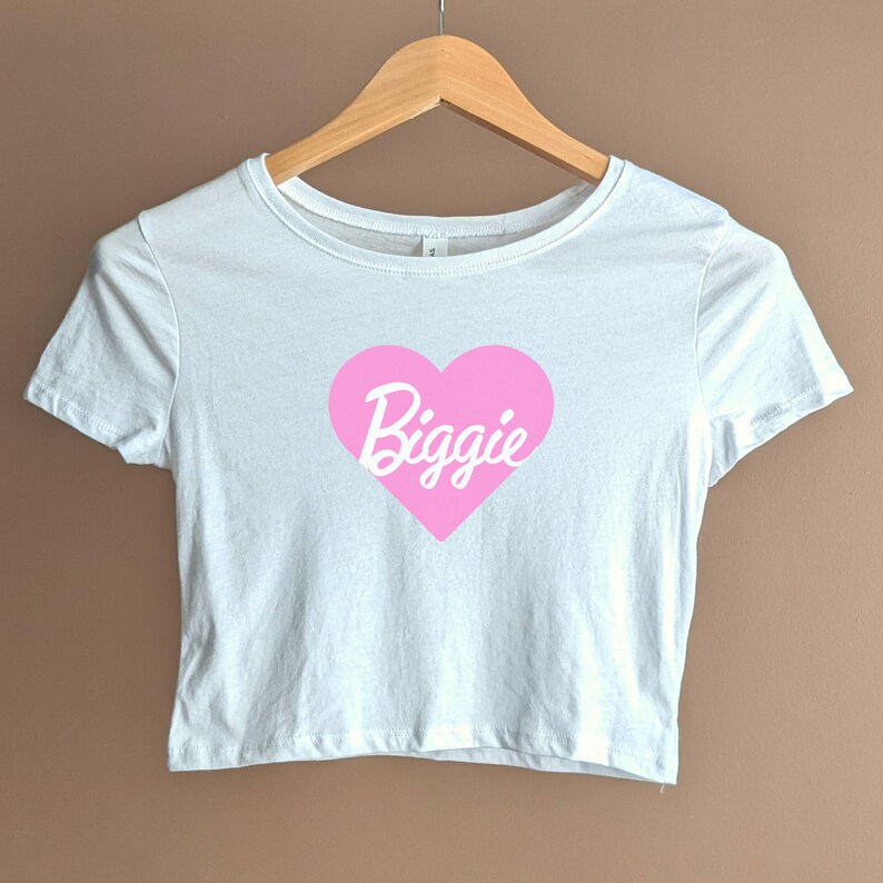 Pu&ograve; includere: Maglietta bianca corta con un grafico a forma di cuore rosa che dice "Biggie" in lettere bianche in corsivo.