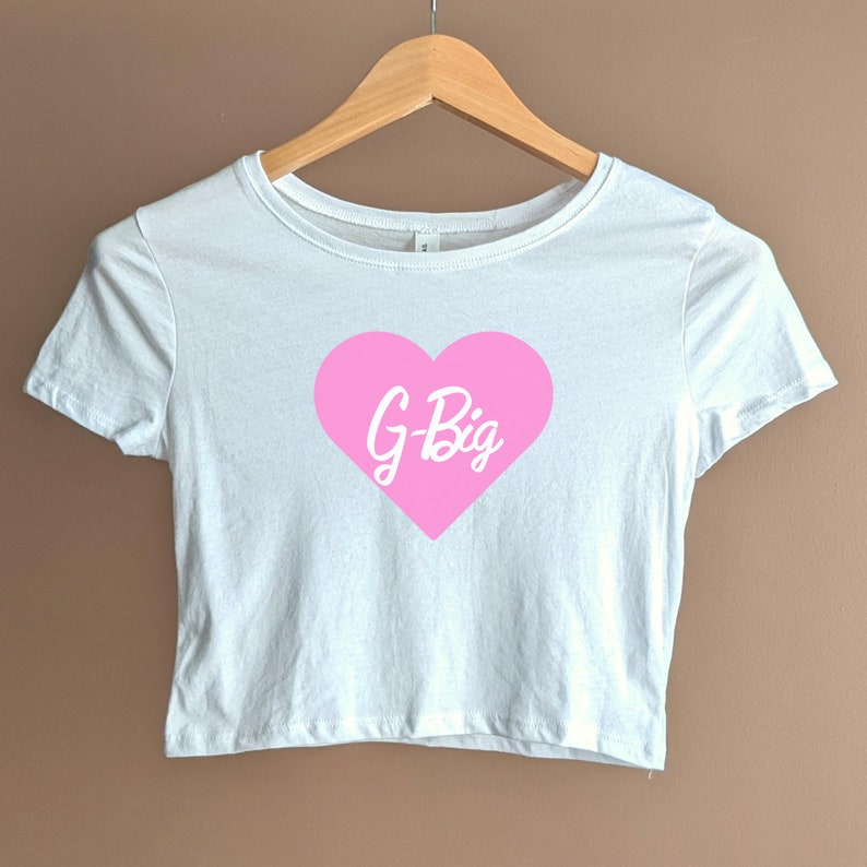 Pu&ograve; includere: Maglietta bianca corta con un grafico a forma di cuore rosa che dice "G-Big" in lettere bianche.