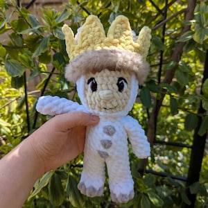 May include: A handmade crochet doll of a character wearing a yellow crown and a furry brown trim. The doll is white with gray accents on the feet and chest. The doll is being held in front of a green leafy background.