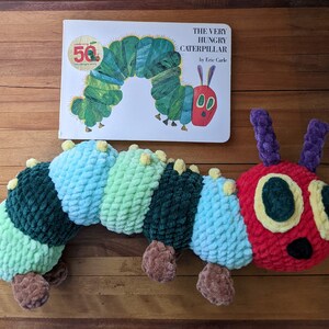 May include: A green and red plush caterpillar toy with a book titled "The Very Hungry Caterpillar" by Eric Carle. The book is celebrating its 50th anniversary.