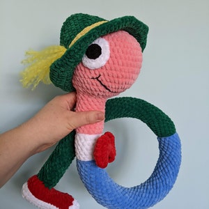 Può includere: Un giocattolo di peluche a forma di verme all'uncinetto che indossa un cappello verde con una fascia gialla. Il verme ha un corpo rosa, braccia verdi e gambe blu e bianche. Il verme tiene un anello verde e blu.
