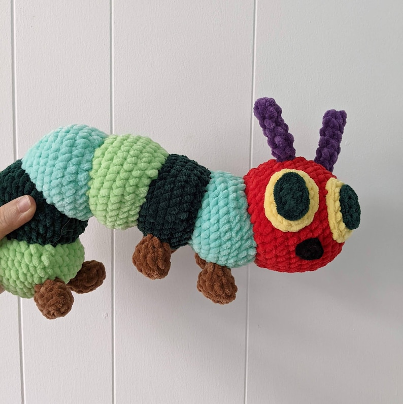 May include: A crocheted plush toy of the very hungry caterpillar. The caterpillar is green, red, and black with a purple and yellow face. The caterpillar is curled in a C shape.