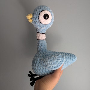May include: A blue crocheted bird plush toy with a white and black collar and large white eyes. The bird has a yellow beak and is standing on a hand.