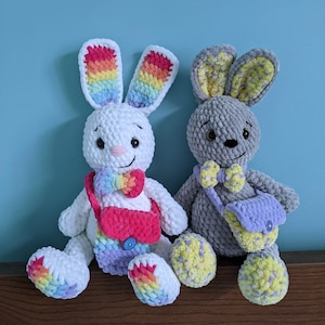 May include: Two crocheted bunny plushies, one white with rainbow ears and a pink bag, the other gray with yellow and purple accents and a purple bag.