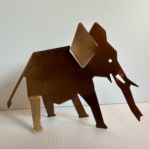May include: A bronze-colored metal elephant sculpture with geometric shapes. The elephant has large ears, a long trunk, and four legs. The sculpture is standing on a white surface. The artist's signature is visible on one leg.