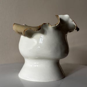 Può includere: Un recipiente in ceramica bianca con un bordo svasato e ondulato caratterizzato da una finitura marrone brunita. L'oggetto presenta una superficie liscia e lucida, un corpo arrotondato e una base ampia e stabile, dalla forma organica.
