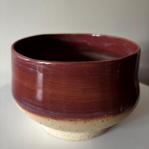 Op de afbeelding: Een keramische kom met een ronde vorm, voorzien van een glanzend, diep roodbruin glazuur op het bovenste gedeelte en een contrasterende, gespikkelde, matte gebroken witte afwerking op de onderkant, wat zorgt voor een tweekleurig uiterlijk.