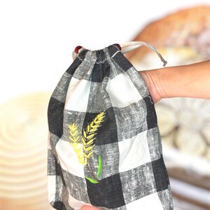 May include: A black and white checkered drawstring bag with an embroidered wheat design in yellow and green. The bag is held in a person's hand, with a loaf of bread visible in the background. The bag has a red bead on each drawstring end.