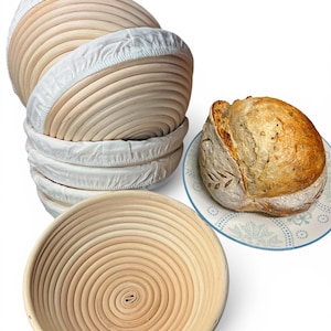 以下が含まれることがあります： 白いライナーが付いた天然木のパン発酵バスケットのコレクション。積み重ねられ、装飾的なプレートに丸いパンと一緒に表示されています。バスケットは、スパイラルデザインの丸型です。