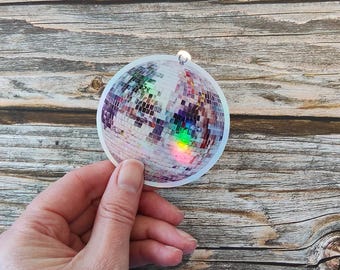 May include: A round, holographic disco ball sticker with a rainbow effect. The sticker is held by a hand against a wooden background. The sticker has a small loop at the top.