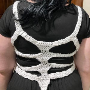 May include: White crocheted skeleton-shaped top with a pointed bottom. The top is worn over a black shirt.