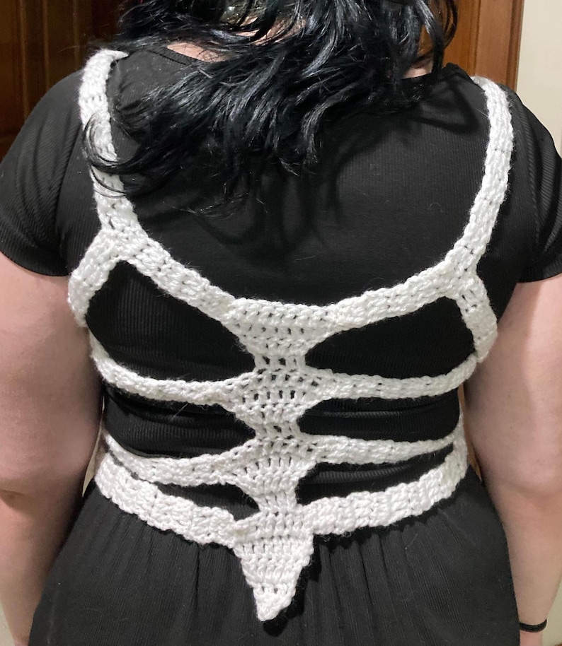May include: White crocheted skeleton-shaped top with a pointed bottom. The top is worn over a black shirt.