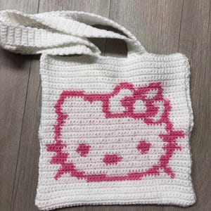 May include: White crochet tote bag featuring a pink Hello Kitty design. The bag has a rectangular shape with a long strap for carrying over the shoulder. The Hello Kitty design is prominently displayed on the front of the bag.