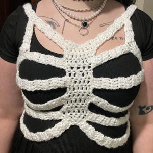 May include: White crocheted rib cage top with a black tank top underneath. The top is worn by a person with a black choker necklace and multiple silver necklaces with pearl accents.