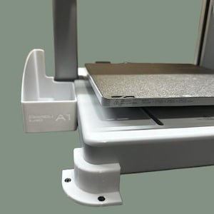 May include: A white and grey 3D printer with a rectangular print bed. A white container with the text "Bambu Lab A1" is attached to the side. The print bed has the dimensions 256x256x256 mm.