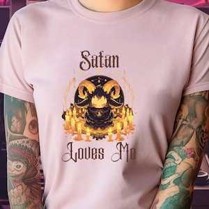 Könnte beinhalten: Ein pinkfarbenes T-Shirt mit einem Motiv einer gehörnten Figur, die von Kerzen umgeben ist, und dem Text "Satan Loves Me".