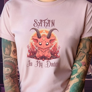 May include: A pink t-shirt featuring a cartoon image of a red devil with horns and the text "Satan Is My Daddy".