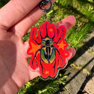 Peut inclure: Un porte-clés en acrylique transparent avec une illustration colorée d'un scarabée. Le scarabée a un corps vert et jaune, avec des accents rouges et est entouré de formes rouges et d'étoiles orange. Le porte-clés a un anneau en métal noir.