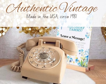 May include: A vintage rotary dial phone in beige with a coiled cord. The phone is sitting on a white surface with a small wooden stand holding a card that says "Happy Birthday Tammy! Leave a Message!" The card has a floral design with blue flowers.