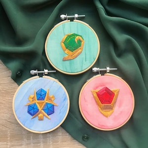 May include: Three embroidered hoops with different colored backgrounds and designs. The first hoop has a green background with a gold and green design. The second hoop has a blue background with a gold and blue design. The third hoop has a pink background with a gold and red design.