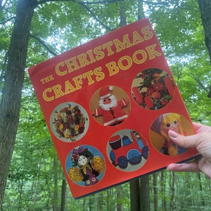 May include: A red book titled "The Christmas Crafts Book" with illustrations of various Christmas crafts, including a Santa Claus, a train, a doll, and a flower arrangement. The book is held in a forest setting.
