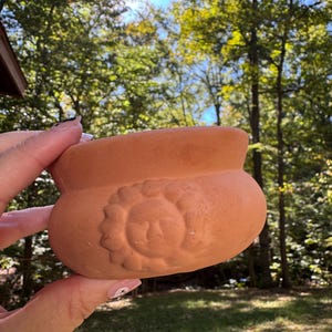 Może przedstawiać: Mała, terakotowa ceramiczna doniczka z motywem twarzy przypominającej słońce. Doniczka ma zaokrąglony kształt z szerokim otworem i lekko podniesioną krawędzią. Tło przedstawia słoneczną scenerię na zewnątrz z drzewami.