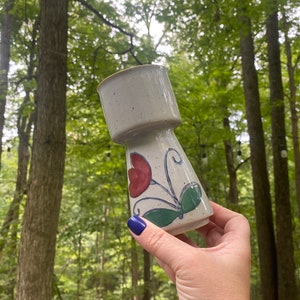 May include: A white ceramic vase with a speckled glaze and a floral design in red, green, and blue. The vase has a wide mouth and a narrow base.