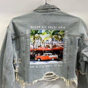 May include: A light blue denim jacket with distressed detailing and a printed image of a vintage car in front of a building with palm trees. The text "Ocean Air Salty Hair", "Beach Drive Good Vibes", "Aire del Oceano Pelo Salado", and "Unidad de Playa Buenas Vibras" is printed on the jacket.