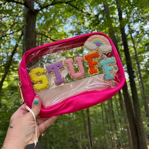 May include: A pink clear plastic makeup bag with a zipper closure. The bag has a rainbow cloud patch and the word "STUFF" in colorful letters.