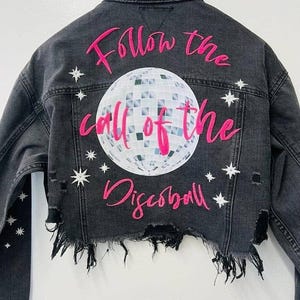 Puede incluir: Una chaqueta vaquera negra con dobladillo deshilachado y estrellas blancas. La parte posterior de la chaqueta presenta un gráfico rosa que dice "Follow the call of the Discoball" con un gráfico de bola de discoteca plateada.