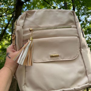 May include: A beige backpack with a gold zipper and a tassel. The backpack has a large front pocket with a gold zipper.
