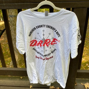 Puede incluir: Camiseta blanca con un estampado gráfico en negro y rojo que dice "Hancock County Sheriff's Dept. DARE Greenfield Police Dept. Being Safe and Responsible".