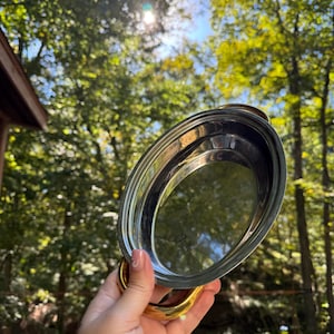Puede incluir: Bandeja ovalada de acero inoxidable con asas doradas. La bandeja tiene una superficie pulida y reflectante. Las asas curvas añaden un elemento decorativo. El fondo es una vista borrosa de árboles verdes y luz solar.