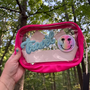 May include: A pink clear plastic zippered pouch with a blue glitter crown patch that says "Travel" and a rainbow smiley face patch with palm trees.