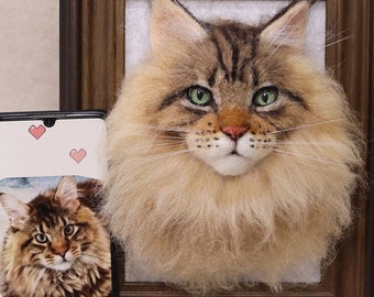 May include: A framed needle felted portrait of a Maine Coon cat with green eyes. The cat has a fluffy brown and white coat. The frame is brown wood with a white mat.