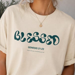 May include: A cream-coloured t-shirt featuring the word "BLESSED" in teal, retro lettering. Underneath is the text "GENESIS 21:22" and a quote. The t-shirt has rolled-up sleeves.