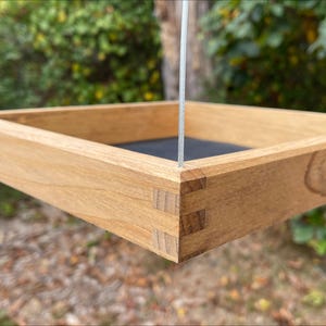 May include: A wooden bird feeder with a square tray and a hanging cord. The feeder is made of light brown wood with a natural finish. The tray is made of a dark material, possibly metal or plastic.