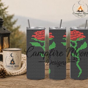 Op de afbeelding: Drie hoge cilindrische bekers met metalen rietjes, voorzien van een zwart honingraatpatroon met een rode roos en groene stengel. Links staat een witte keramische mok met de tekst Campfire Moon Designs en een kampvuurafbeelding.